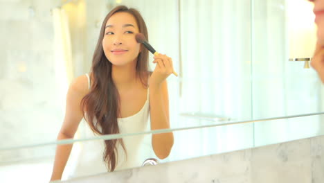 A-pretty-young-woman-with-flawless-skin-looks-in-the-mirror-as-she-applies-makeup-with-a-large-blush-brush