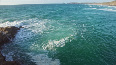 View-of-Foamy-Turquoise-Green-Waves-Splashing-Over-Coastal-Rocks-from-Cliffside,-Cornwall---slow-motion