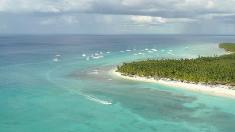 A-large-group-of-luxury-yachts-anchored-just-offshore-as-powerboat-cruises,-Saona,-aerial-panorama