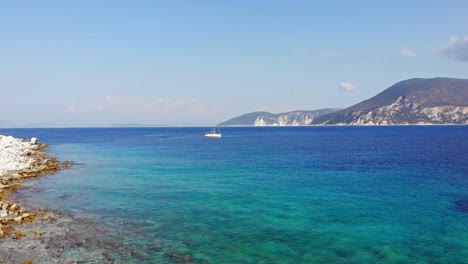 Rocky-Shoreline-Of-Emplisi-Beach-With-Sailing-Boat-At-Summer-In-Greece