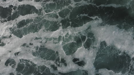 A-top-down-drone-shot-of-white-froth-and-waves-rolling-by-in-the-sun