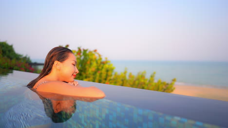 A-close-up-of-a-beautiful-woman-at-the-edge-of-a-swimming-pool-leans-her-arms-along-the-edge-as-she-looks-out-at-the-ocean-view