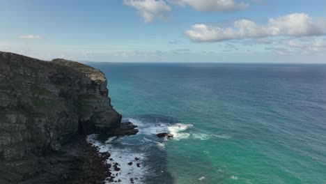 Aerial-reveal-shot-flying-along-rugged-cliffs-by-a-beautiful-turquoise-ocean-on-a-sunny-day
