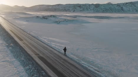Caminante-Solitario-En-Una-Carretera-Remota-En-Un-Paisaje-Nevado-De-Invierno-En-Islandia,-Aéreo