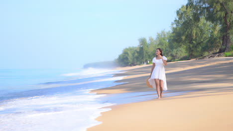 A-young-fit-woman-in-a-white-sundress-walks-along-the-beach-while-the-waves-roll-in