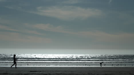 Silhouetten-Von-Menschen-Am-Strand---Ein-Spazierendes-Mädchen-Und-Ein-Kind,-Das-In-Zeitlupe-Nach-Muscheln-Sucht