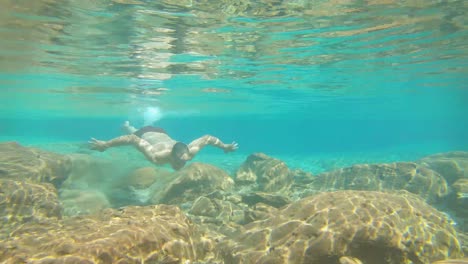 Junger-Mann-Schwimmt-In-Blau-Klar-Unter-Wasser-Des-Wasserfalls-Mit-Vielen-Felsen-Aus-Flachem-Winkel
