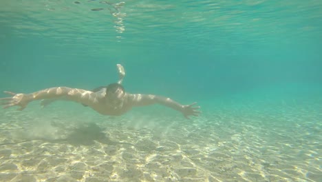 young-man-swimming-in-blue-clear-under-water-of-waterfall-with-many-rocks-from-flat-angle