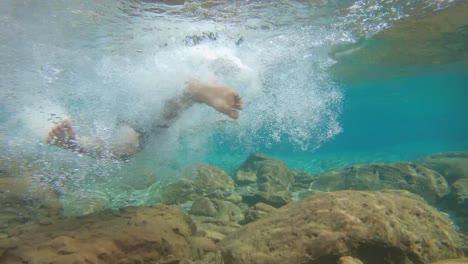 Junger-Mann-Schwimmt-In-Blau-Klar-Unter-Wasser-Des-Wasserfalls-Mit-Vielen-Felsen-Aus-Flachem-Winkel