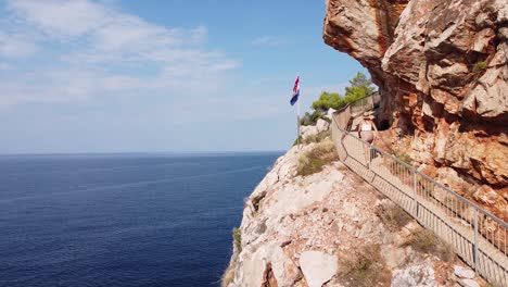 Tourist-Girl-Walks-along-Steep-Cliff-at-Pasjaca-Beach,-Dalmatia,-Croatia---Aerial-Dolly