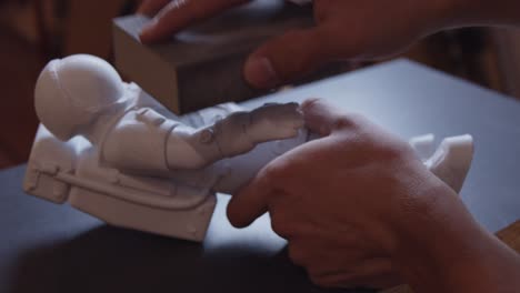 Sanding-Hand-of-a-Small-White-Astronaut-with-Sand-Paper