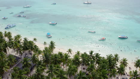 Luxury-yachts-and-crowds-of-tourists-gather-along-white-sand-beaches-of-Saona-Island,-aerial-reveal