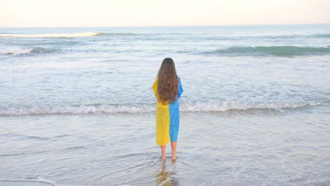 Junges-Mädchen-Mit-Lockigen-Blonden-Haaren,-Das-Eine-Ukraine-Flagge-Auf-Dem-Rücken-Trägt-Und-Meereswellen-Beobachtet,-Die-An-Die-Küste-Rollen