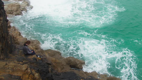 Lone-Teenager-Seated-on-Cliffside-Rocks-as-Turquoise-Ocean-Waters-Crash-Against-Rocks---looking-down-reveal-shot