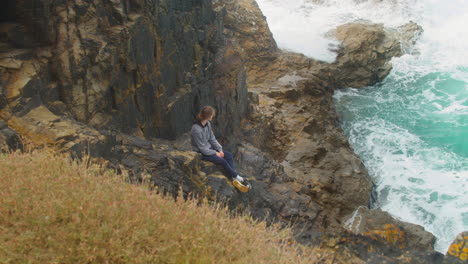 Teen-Lad-Sitting-on-Coastal-Cliff-Edge-Watching-Turquoise-Ocean-Waves-Crash-Over-Rocks,-Cornwall,-England---Static-Shot