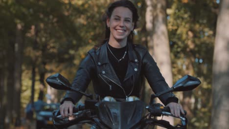Beautiful-closeup-of-smiling-white-Female-on-motor-bike-driving-in-leather-jacket-riding-in-forest,-trees-with-golden-autumn-leaves-color-on-sunny-day