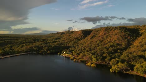 Beautiful-drone-shot-of-green-growing-hills-beside-water-lake-in-the-evening