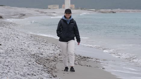 Happy-woman-walking-alone-on-a-beach-in-Ibiza,-wearing-a-jacket-and-cap---slow-motion-shot
