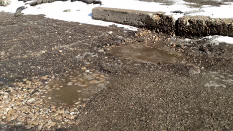 Moving-along-a-broken-road-with-snow-on-the-edges-and-potholes-filled-with-water-and-rocks