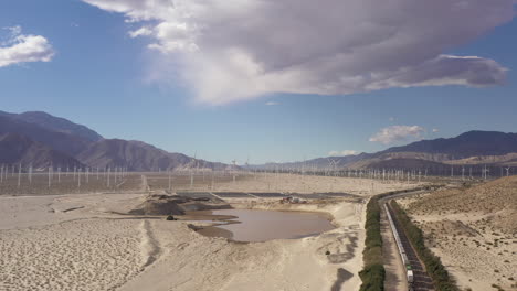 Aerial-of-cargo-train-coming-through-near-Palm-Springs-Wind-Farm,-mountains-in-distance