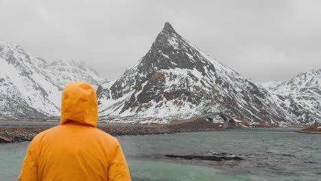 Man-In-Yellow-Hooded-Jacket-Admiring-Famous-Bridge-And-Beautiful-Snowy-Landscape-Of-The-Lofoten-Islands---wide-shot