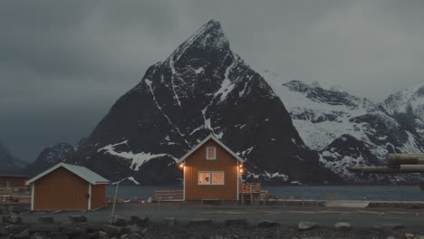 Picturesque-View-Of-A-Lighted-Small-Cabin-With-Background-Of-Olstinden-Mountain-At-Twilight-In-The-Village-of-Sakrisoy,-Lofoten-Islands-Norway---wide-shot