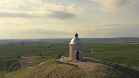 A-newlywed-couple-walking-holding-hands-around-a-small-chapel-on-a-hill-overlooking-vineyards,-aerial-orbit