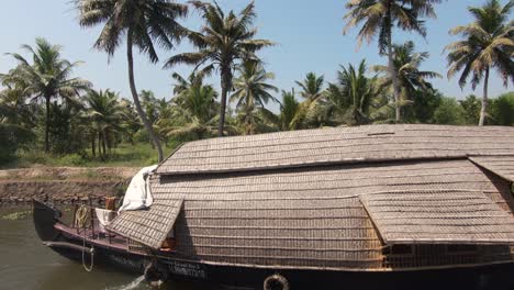 Palm-tree-river-shore-in-Alappuzha,-wooden-houseboat-sailing-in-waterway