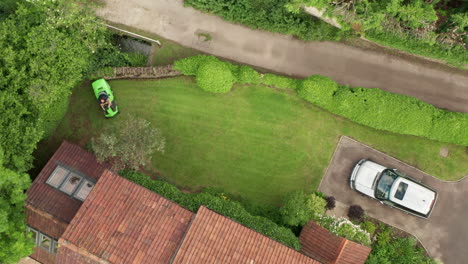 Rising-Aerial-Shot-of-Man-Mowing-Lawn-of-Home-on-Summer’s-Day-using-Ride-On-Lawnmower-from-Birds-Eye-View-Perspective
