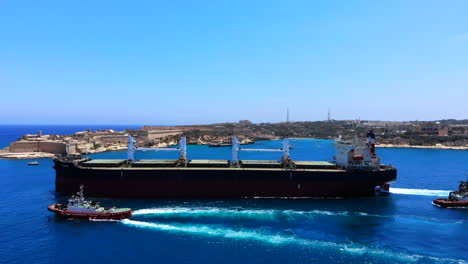Aerial-shot-of-an-enormous-cargo-ship-leaving-the-harbour-of-Valletta,-deep-blue-sea,-tug-boats,-old-city-in-the-background