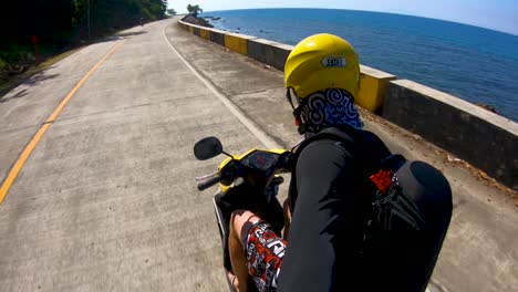 A-smooth-panning-shot-of-a-man-riding-a-motorcycle-along-a-beautiful-sunny-coastal-road-on-the-island-of-Bohol,-Philippines