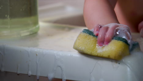 Cute-toddler-sitting-in-highchair-cleaning-with-soapy-sponge-and-water-on-white-table-top