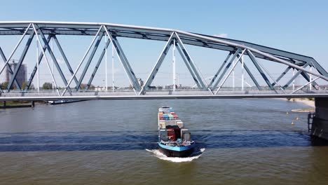 Container-Ship-Sails-Under-Bridge-at-Waal-river-in-Nijmegen,-Gelderland,-Netherlands---Aerial-Pan-Left