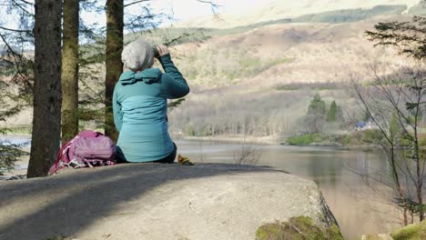 A-woman-sits-on-a-rock-eating-her-lunch-and-drinking-tea-while-out-for-a-walk-in-the-forest,-looking-out-across-a-Scottish-Loch-and-hillside-in-the-sun