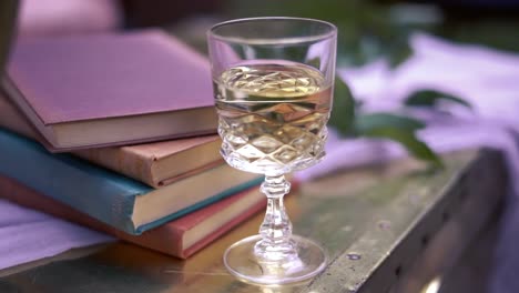 A-rearward-bokeh-shot-of-a-diamond-styled-glass-of-white-wine-beside-a-stack-of-colorful-books-and-leaves-on-top-of-a-white-cloth-on-a-wooden-table