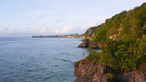 Beautiful-rotating-aerial-shot-of-the-exotic-Padang-Padang-beach-with-a-woman-standing-on-the-edge-of-a-cliff