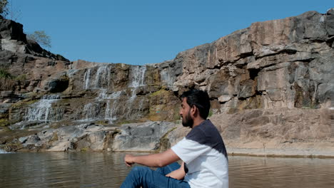 Bearded-man-sitting-near-waterfall-throwing-stone-into-river