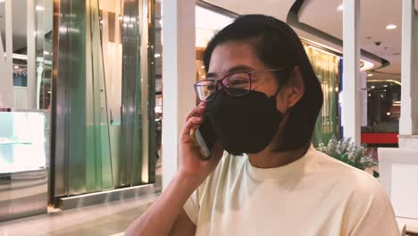 Portrait-Asian-middle-aged-woman-talking-with-a-mobile-phone,-wearing-a-face-mask,-being-in-shopping-mall