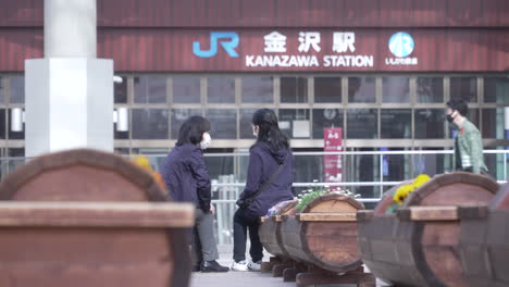 Two-Women-Wearing-Masks-Sitting-And-Chatting-In-Front-Of-The-Entrance-Of-Kanazawa-JR-Train-Station-During-Pandemic-In-Japan