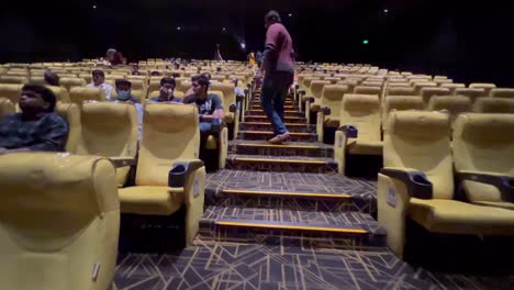 There-are-several-individuals-seated-in-their-seats-in-the-cinema-hall-or-movie-theatre-as-the-person-wearing-the-purple-shirt-ascends-the-steps-to-his-seat