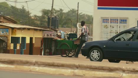 Slowmotion-Shot-of-Street-Coffee-Vendor-Pushing-His-Cart-with-Cars-and-Motorcycles-Passing-in-Foreground---Lomé,-Togo,-Africa