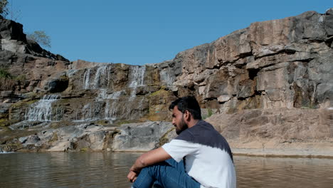 Tired-bearded-man-sitting-by-river-near-waterfall-in-sunny-day