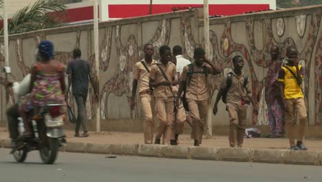 Afrikanische-Studenten-Zu-Fuß-Am-Straßenrand-Mit-Vorbeifahrenden-Autos-Und-Motorrädern-Im-Vordergrund---Togo,-Afrika
