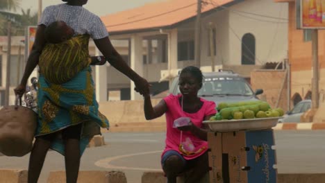 Slow-mo-4K-shot-of-African-female-water-street-vendor-carrying-a-baby-in-the-back-and-trading-with-a-fruit-vendor,-Togo,-Africa
