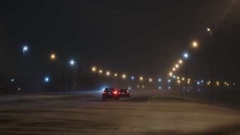 Static-shot-of-cars-driving-on-a-well-lit-highway-during-a-blizzard-with-loose-snow-on-the-road-being-blown-by-strong-winds-at-night