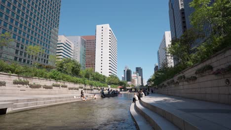 People-resting-sitting-by-Cheonggyecheon-Stream-on-a-Summer-hot-Day-in-Seoul-City-downtown-with-a-view-of-high-towering-skyscrapers,-South-Korea