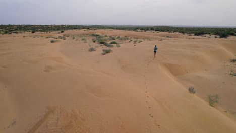Scenic-drone-aerial-view-tracking-a-young-man-running-through-dunes-in-the-desert