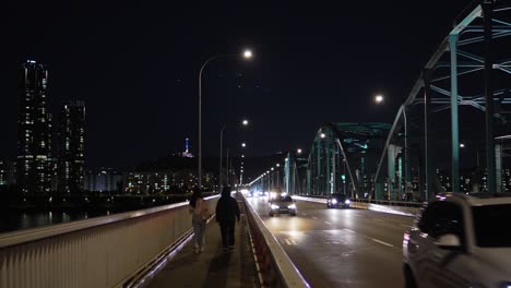 Seoul-nightlife---Back-of-two-young-Korean-girls-walking-on-Dongjak-bridge-at-night-roadside-of-a-multilane-freeway-with-car-traffic,-skyscrapers-in-the-background