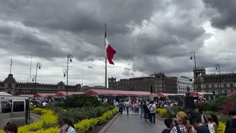 Toma-De-La-Bandera-De-México-Ondeando-Con-La-Vista-Completa-Del-Zócalo-En-El-Fondo-Al-Atardecer-Durante-Una-Feria-Artesanal