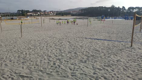 Adult-beach-soccer-coach-with-children-watches-the-activity-among-the-volleyball-nets-in-brightly-colored-T-shirts-one-summer-morning,-rolling-shot-to-the-right
